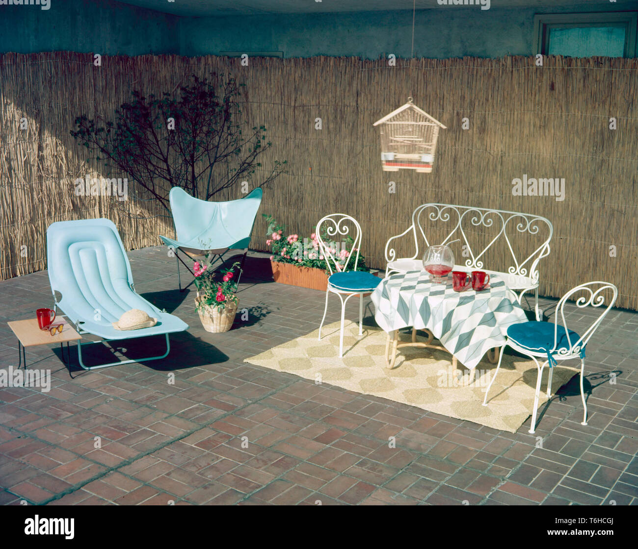 Home decor of the 1950s. A set of matching garden furniture with blue cushions. The chair is the typical Butterfly chair type, also known as a BKF or Hardoy chair. There is also a matching set of white painted iron furniture with two chairs and a sofa. The table is set with a bowl of lemonade and two glasses. Stock Photo