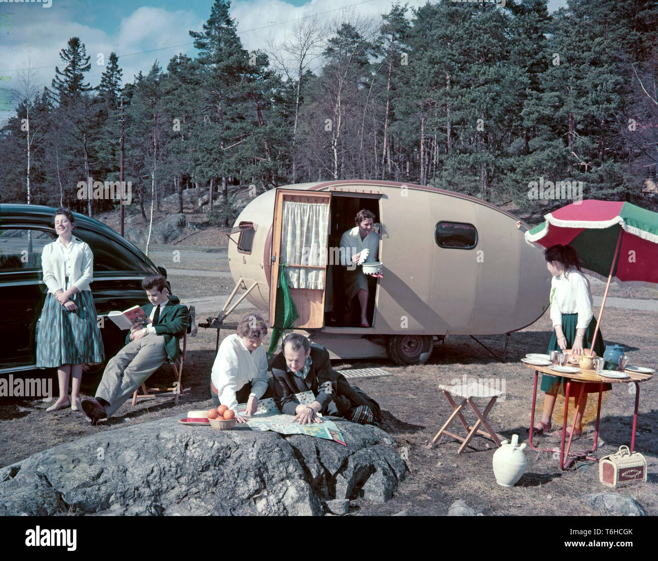1950s camping. A family is enjoying their holiday and the practial camping life in their caravan. Demonstrating how well everything works for them even on holiday. The mother holds out a pot with a ready cooked meal while her daughter has set the table. The father and daughter spends their time looking at a map probably to find the next suitable location and campsite. Everything in this pictures illustrates very well the time of the 1950s both in visible objects and clothing. Other aspects of the time is that while the women is pictured cooking, the man is not. Note the model of the caravan an Stock Photo