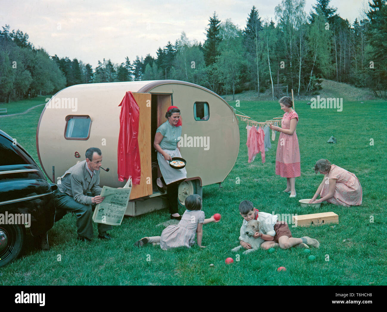 1950s camping. A family is enjoying their holiday and the practial camping life in their caravan. Demonstrating how well everything works for them even on holiday. The mother has fried some eggs and and steps out of the caravan holding the pan. Her eldest daughters are washing dishes and hanging up the laundry to dry. The youngest children are playing and the father of course sits around doing nothing more then having a smoke and reading the newspaper as a typical 1950s father would do.Everything in this pictures illustrates very well the time of the 1950s both in visible objects and clothing. Stock Photo