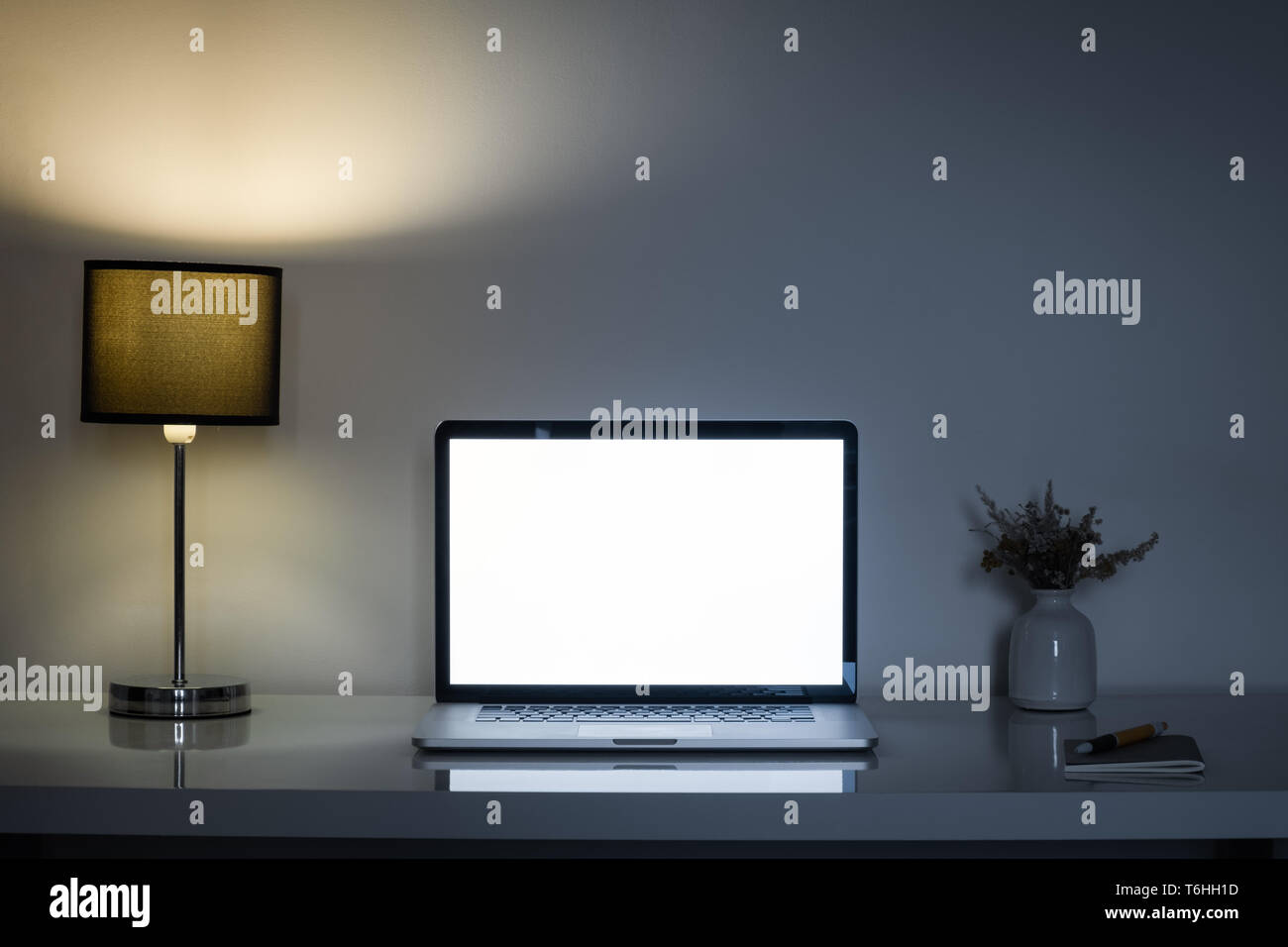 Cozy home office desk at night lit by a lamp and white laptop screen. Clean modern workplace for telework or freelance activity, low key mood Stock Photo