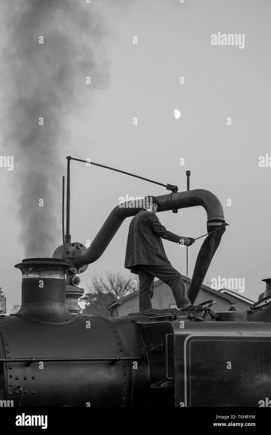 Black & white shot of steam locomotive crew member standing isolated on top of vintage UK steam engine using water crane and filling up water tank. Stock Photo