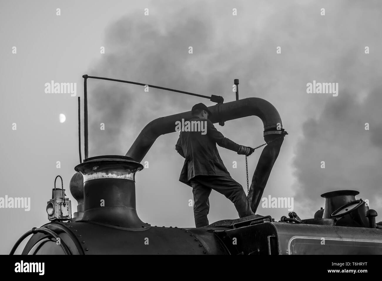 Black & white shot of steam locomotive crew member standing isolated on top of vintage UK steam engine using water crane and filling up water tank. Stock Photo