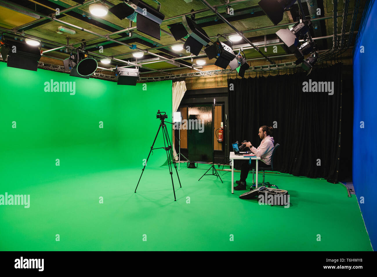 A wide-angle shot of a caucasian man working at his desk in a film studio, a green screen surrounds the room interior, ready for use. Stock Photo