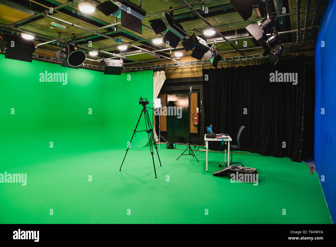 A wide-angle shot of a film studio, a green screen surrounds the room interior, a tripod stands in the middle of the room with a camera ready to film  Stock Photo