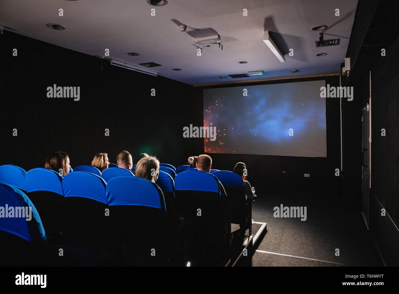A rear view shot of a movie theater, unrecognizable people can be seen sitting together in blue seats in a row infront of the projection screen. Stock Photo