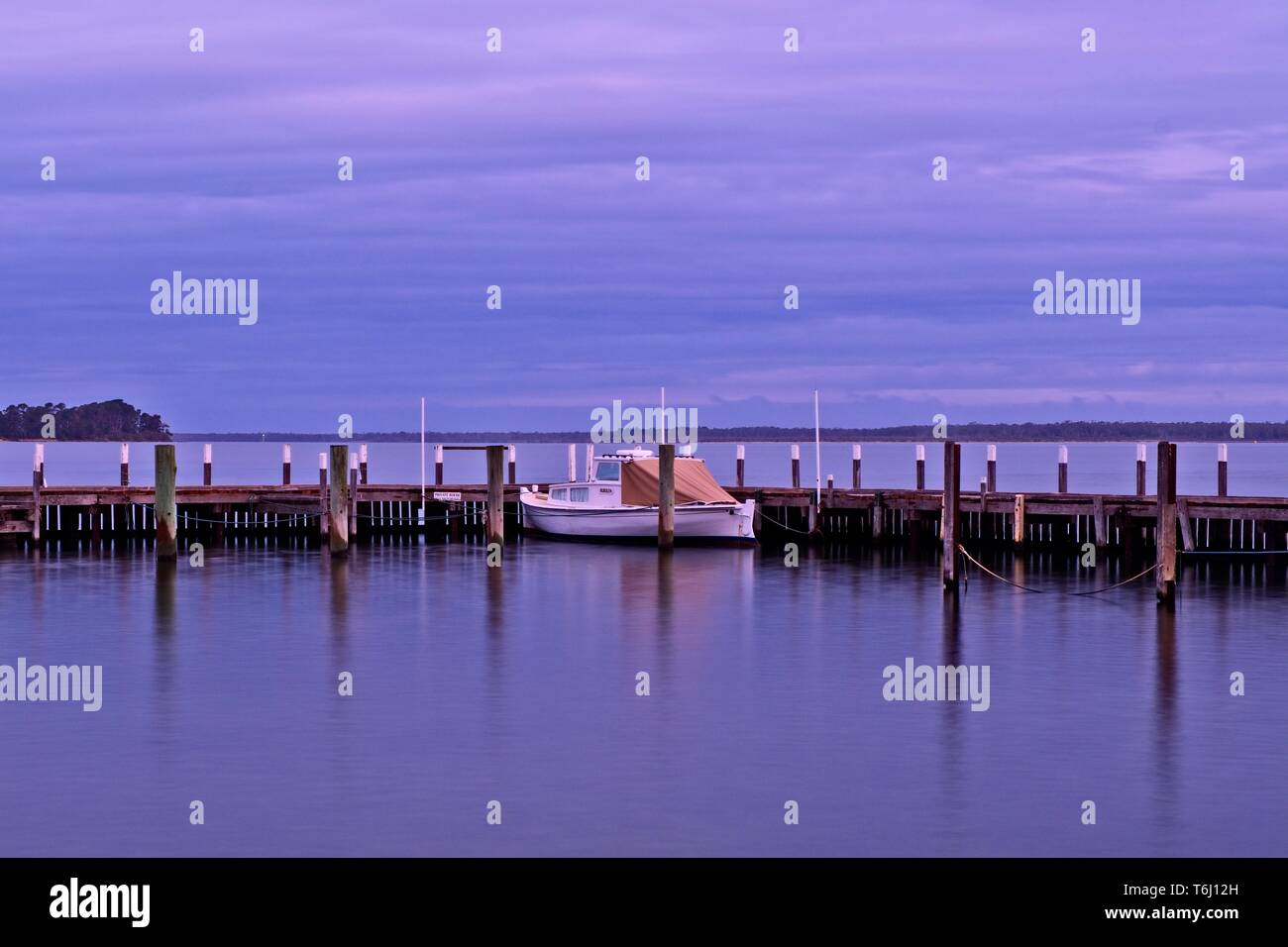 The calm of mauve colours in a twilight shot of a small boat moored at a rustic wooden jetty in Metung with Gippsland Lakes in the background Stock Photo