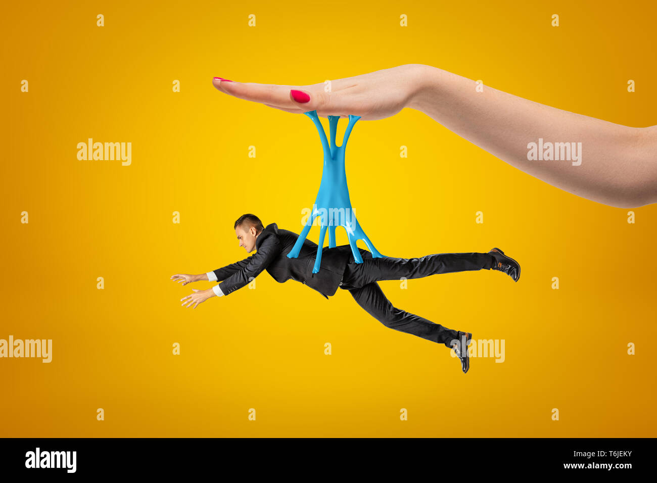 Side view of little man in suit hanging on blue sticky slime stuck to big woman's hand above on yellow background. Stock Photo
