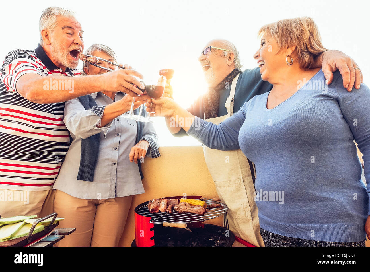 Happy senior friends having fun cheering with red wine at barbecue in terrace outdoor - Mature people dining and laughing together on rooftop Stock Photo