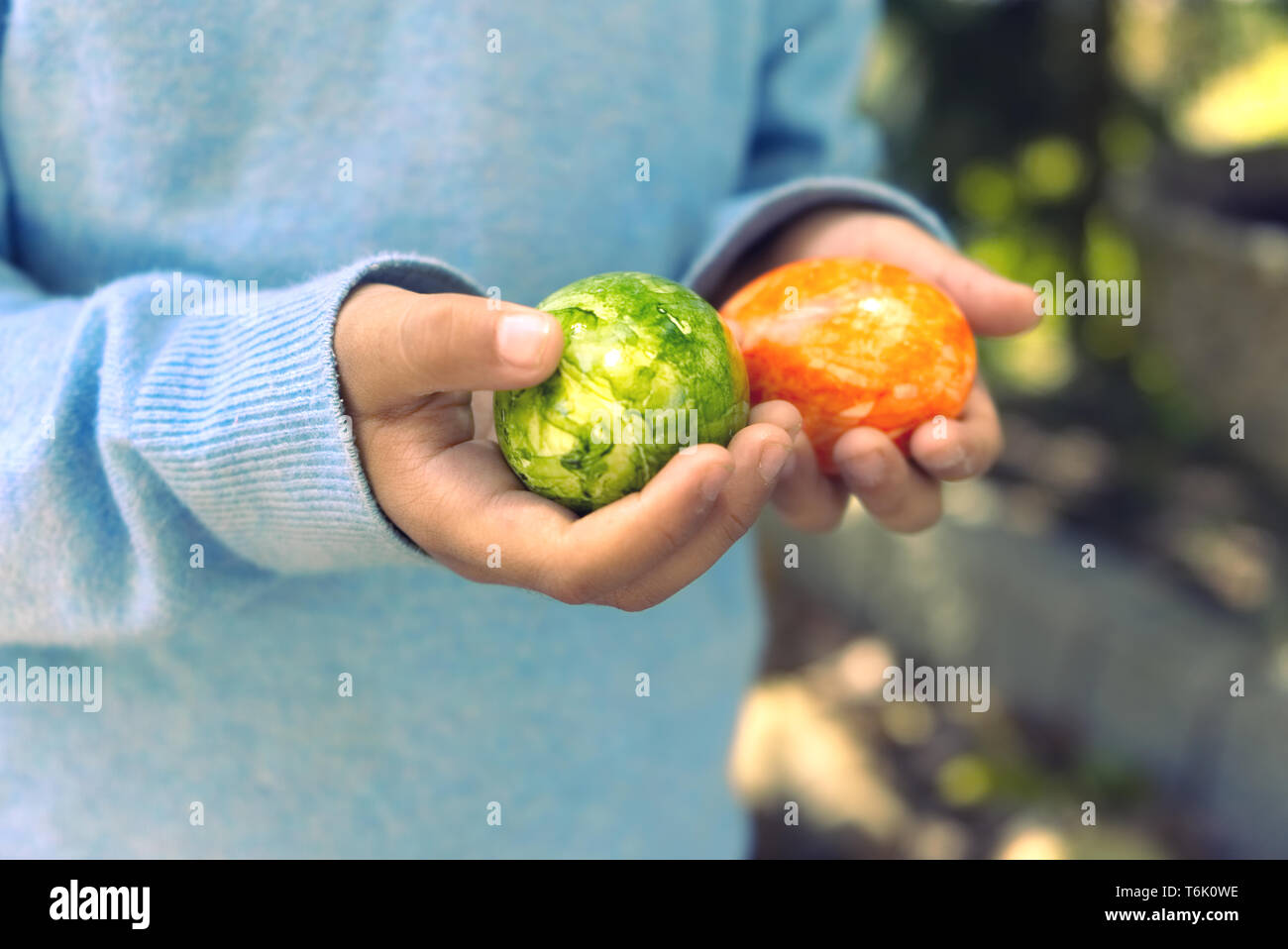 Mid section of a child with light blue sweater holding two green and orange painted ester eggs in an outdoor setting. Stock Photo