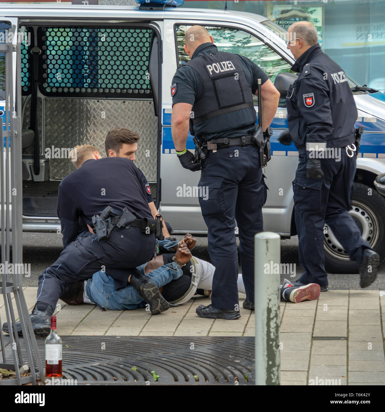 Wolfsburg, Germany, May 1, 2019: A dark-skinned man is handcuffed by German police officers in black uniforms and held at the bottom of the sidewalk,  Stock Photo