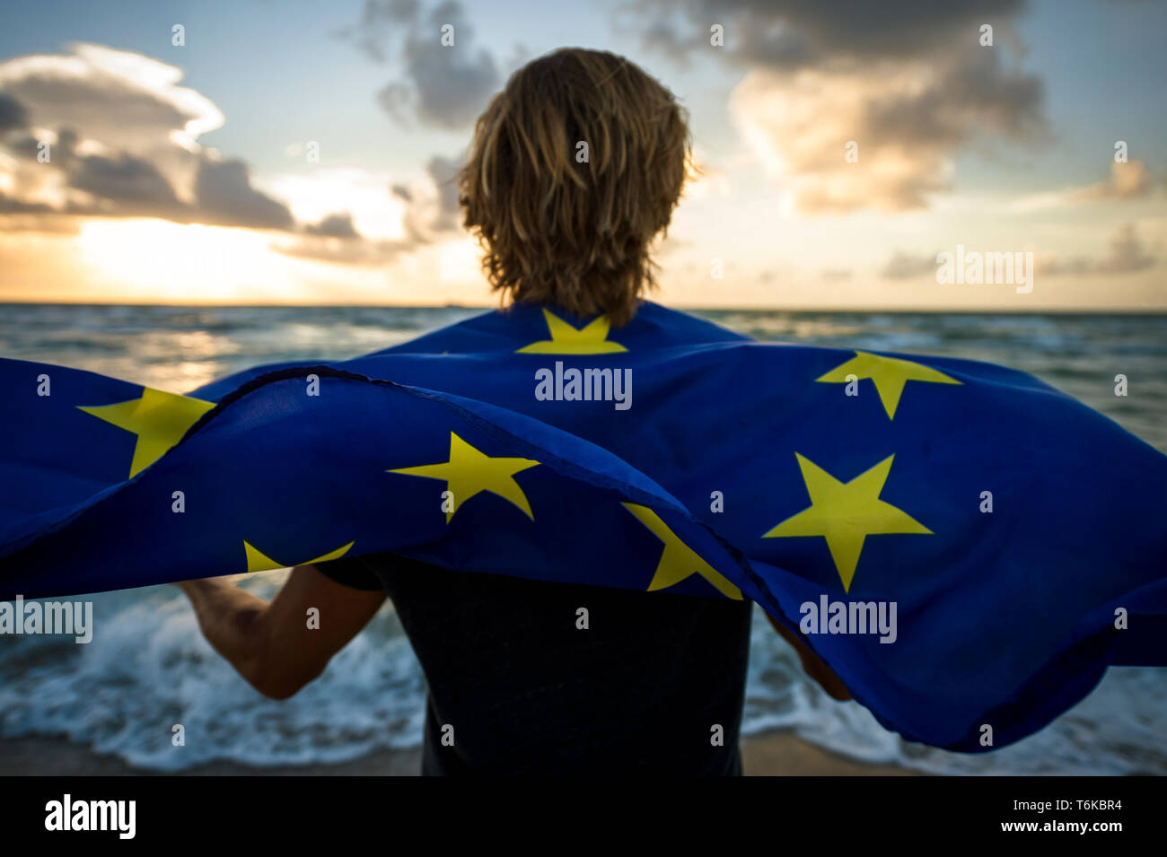 Man with blond hair holding a fluttering EU European Union flag on a sunrise beach  in front of stormy turbulent Channel seas Stock Photo