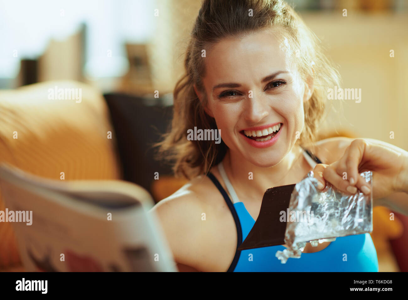 Portrait of happy fit sports woman in sport clothes in the modern house reading magazine and eating chocolate. Stock Photo