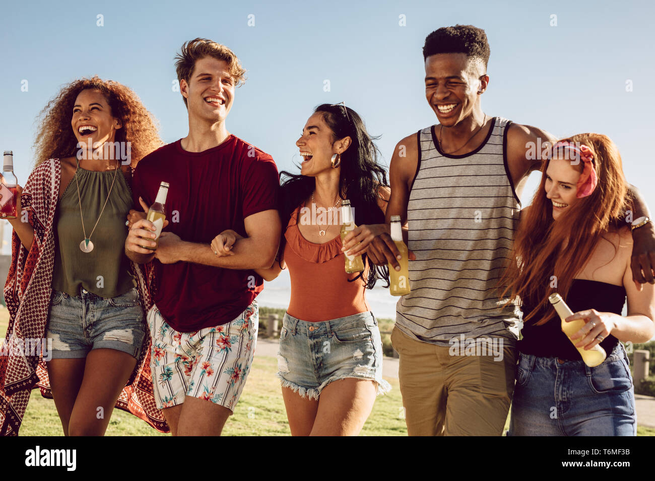Group of friends walking together with locking arms. Young men and women with beers walking together and having fun. Stock Photo
