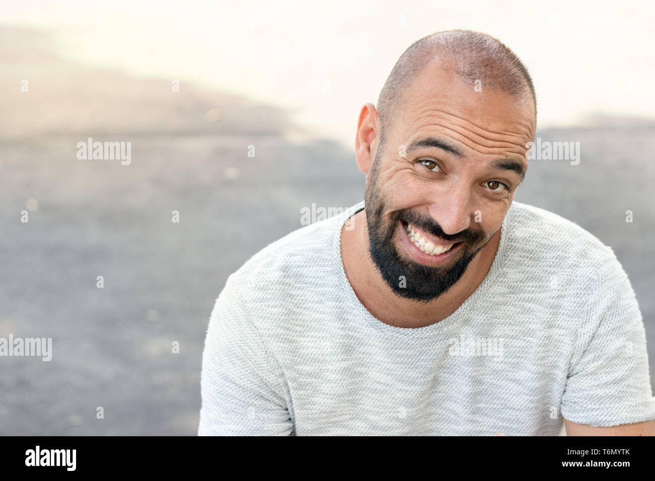 Portrait of a spanish man with a beard and ultra-short buzz, smiling in a funny way, looking camera, wearing a t-shirt, outdoors. Stock Photo