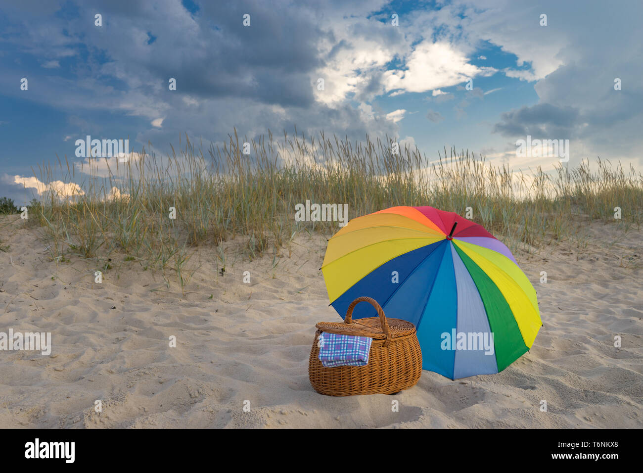 Multicolored umbrella and picnic basket against wild beach and clouds, weekend break concept Stock Photo