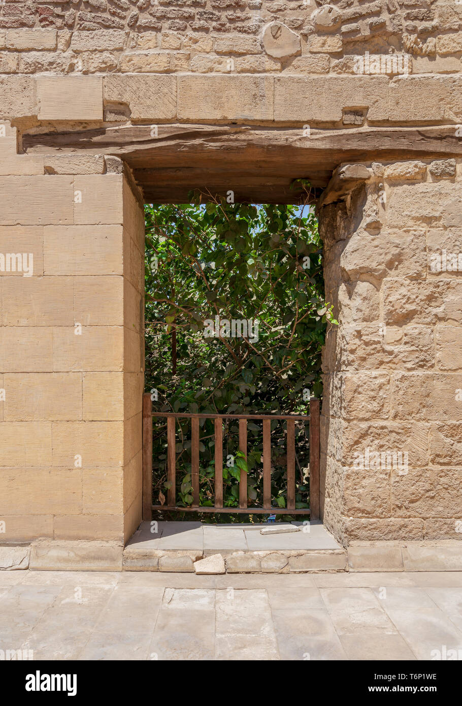 Rectangular opening with wooden balustrade on old abandoned stone bricks wall revealing part of a big green dense tree in summer time Stock Photo