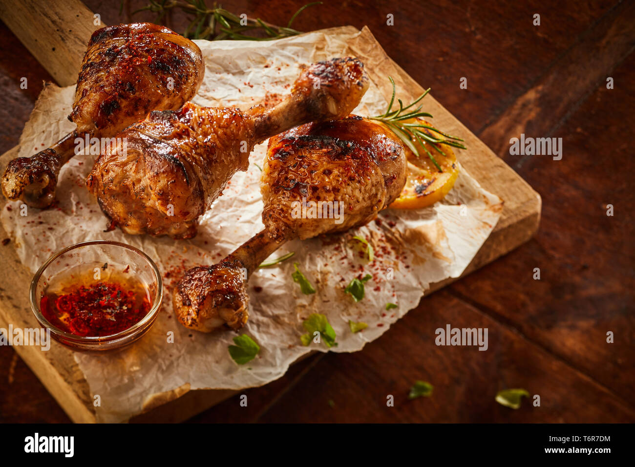 Spicy grilled chicken drumsticks with chili sauce or dip and fresh herbs served on paper on a wooden chopping board Stock Photo