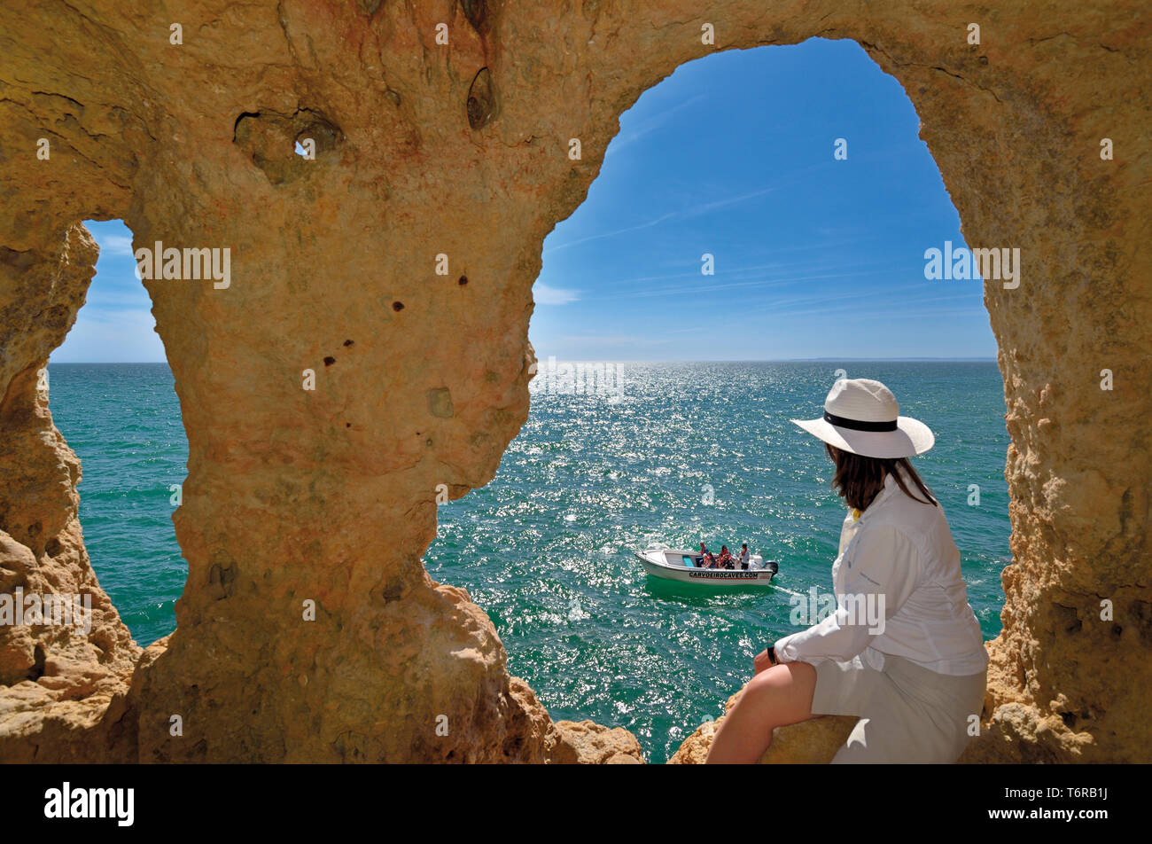Woman looking from natural cave window to green ocean and  passing touristic boat on a sunny day Stock Photo