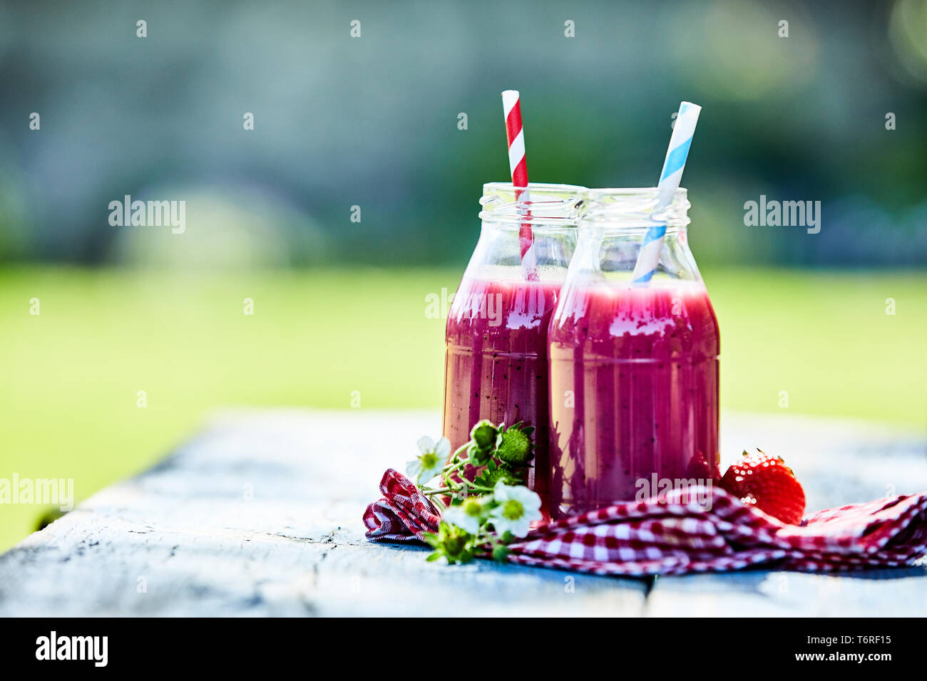 Bright, vibrant strawberry smoothies in glass jars on an outdoor summer picnic table setting. Stock Photo