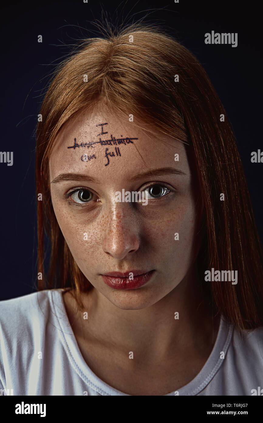 Portrait of young woman with mental health problems. Tattoo on the forehead with the words I have bulimia-I'm full. Concept of hidding the true feelings, psycological trouble, treatment. Stock Photo