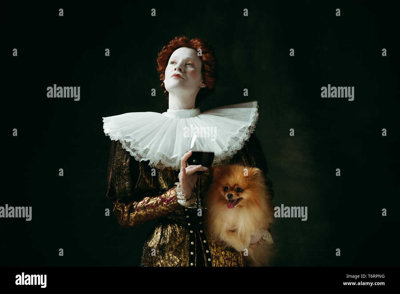 Feel right. Medieval redhead young woman in golden vintage clothing as a duchess holding puppy and glass with red wine on dark green background. Concept of comparison of eras, modernity, renaissance. Stock Photo