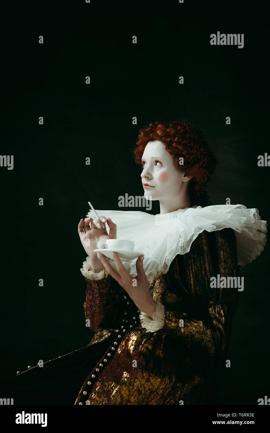 Medieval redhead young woman in golden vintage clothing as a duchess holding a cigarette and a cup of coffee on dark green background. Concept of comparison of eras, modernity and renaissance. Stock Photo