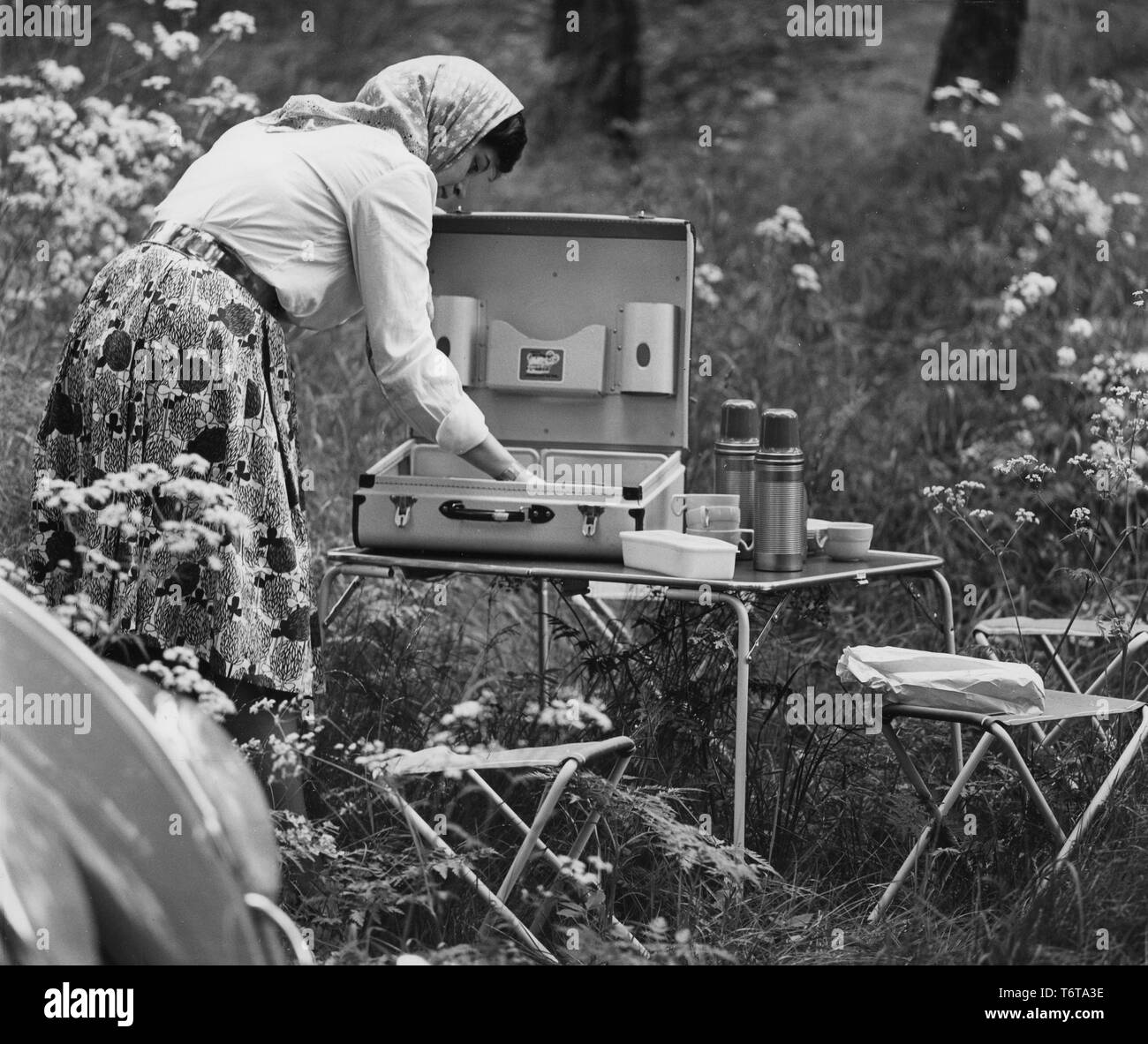 Camping in the 1960s. When camping in the 1960s plastic cups and other camping equipment were sometimes packed and transported in special cases. Pictured a practical such and a woman setting the table for a meal in the nature. The chairs and the table are typical camping folding ones, that could easily be stored and transported, not to take up too much room in the trunk of the car. Sweden 1962 Stock Photo