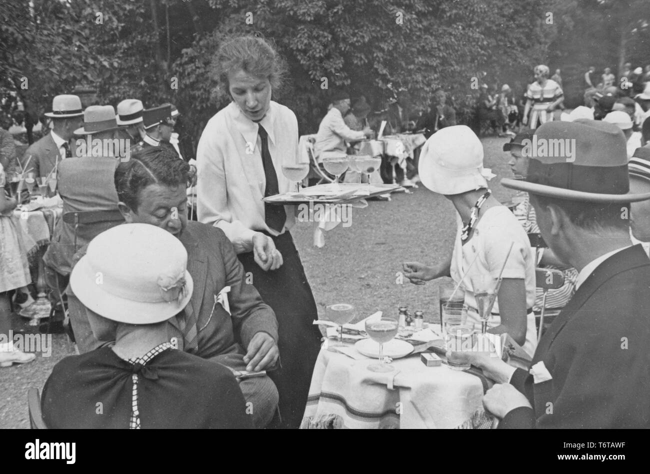 1930s open-air cafe. It's full of people eating and drinking. A female waitress is both serving drinks and receives payments from the customers. Sweden 1930s Stock Photo