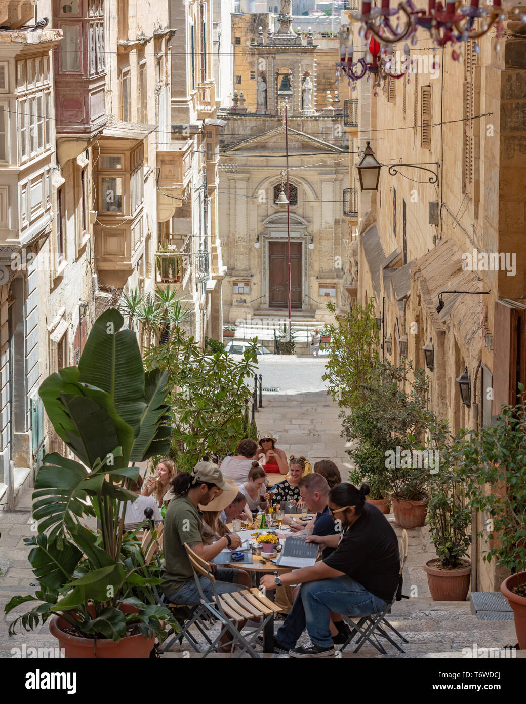 Friends enjoy an al fresco lunch at a restaurant in the narrow St. Lucy's Street in Valletta, with the Church of St Lucy in the background. Stock Photo