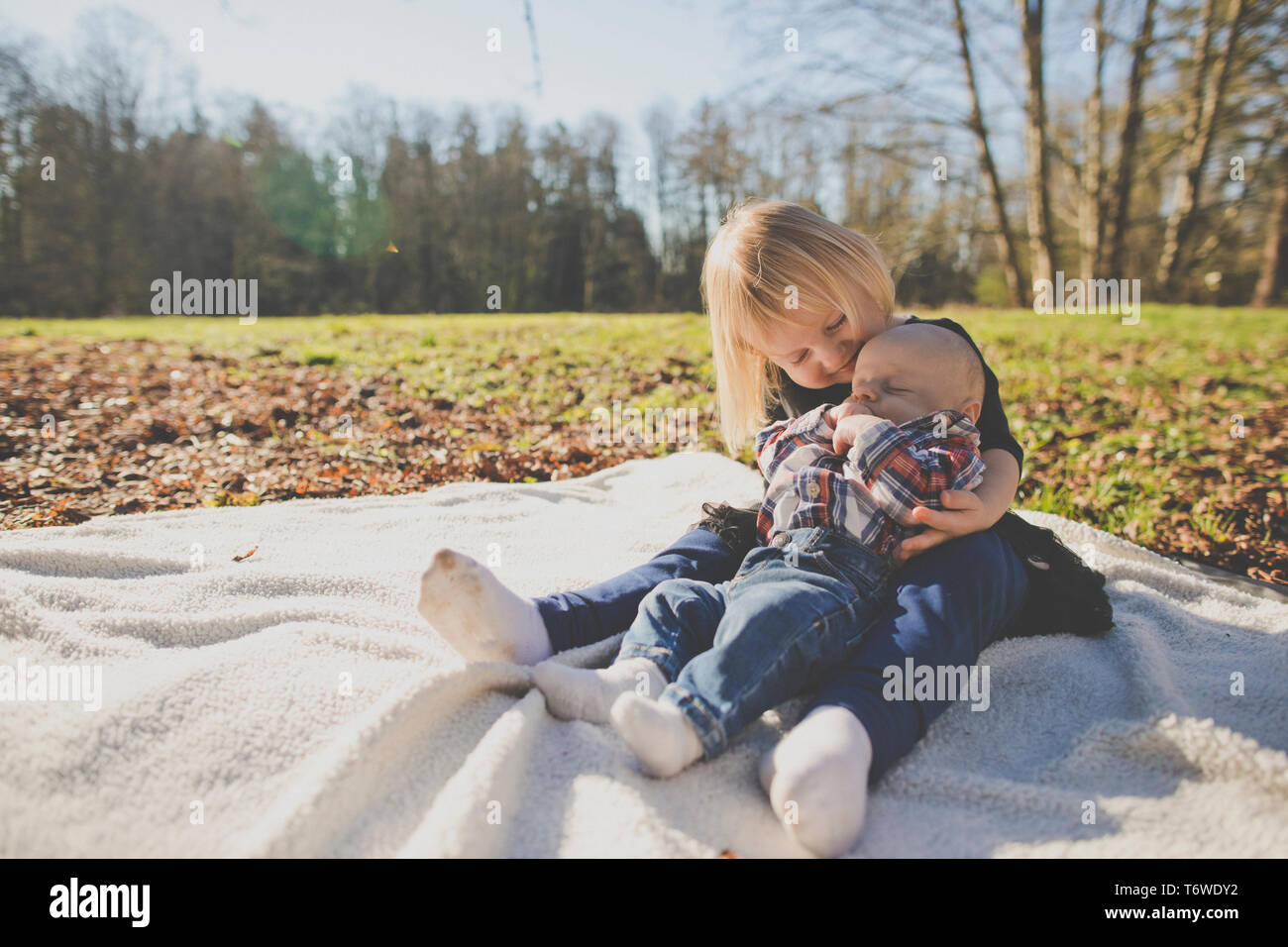 Sister holds her brother while on picnic blanket outside at park Stock Photo