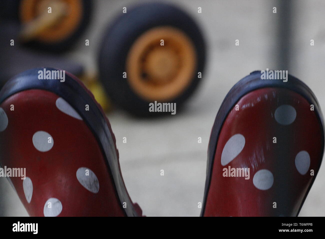 colour image of a close up view of a pair of red childrens boots with white dots. in the background is a yellow tyre from a toy. Stock Photo