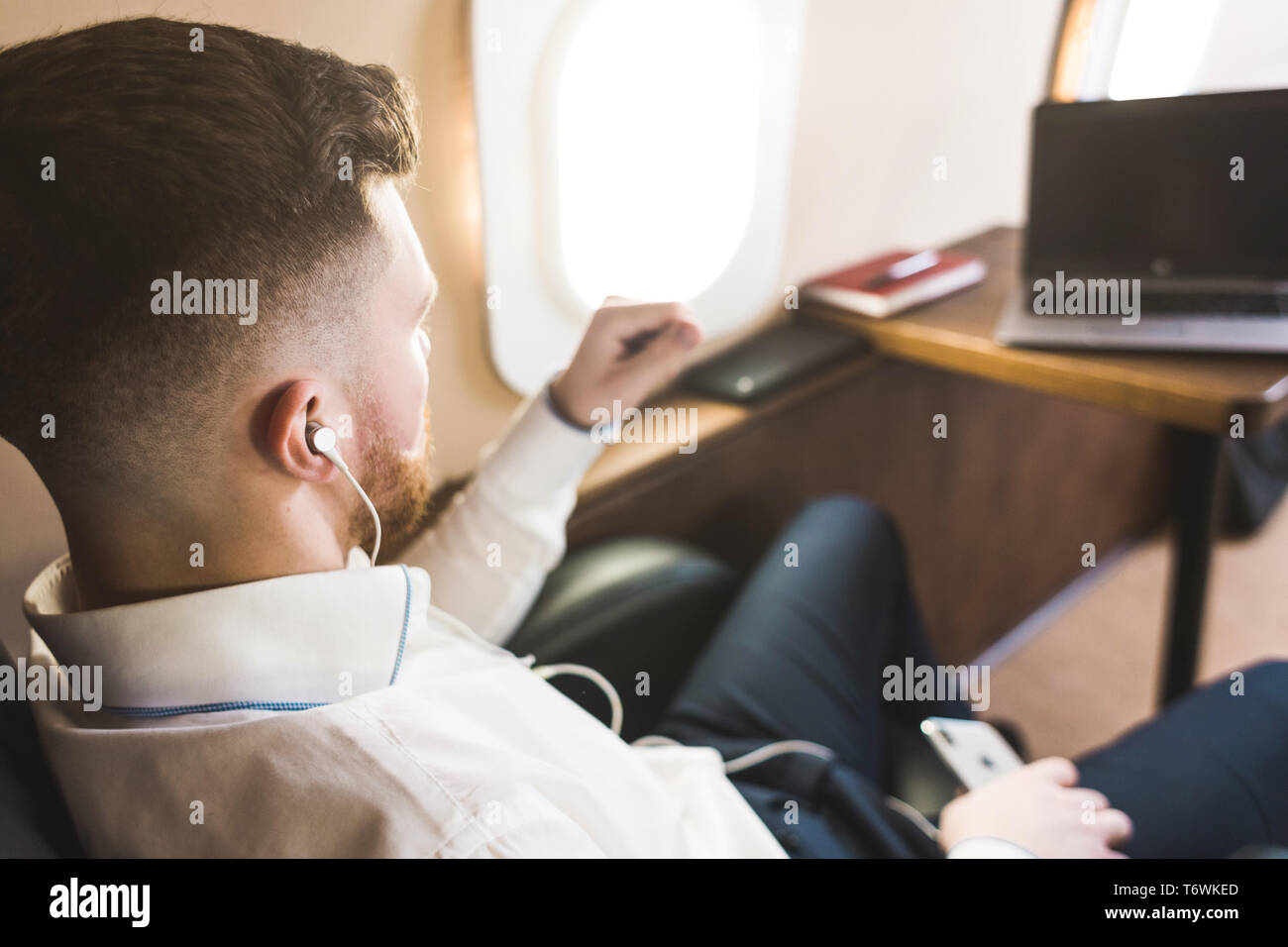 Young attractive and successful businessman is resting and listening to music works while sitting in the chair of his private business plane Stock Photo