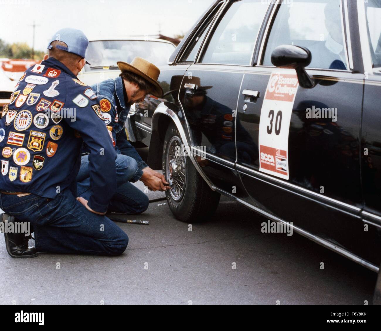 Tow men, wearing jackets with patches, check the tire of a black sedan after a competitive fuel efficiency driving event, 1975. Image courtesy US Department of Energy. () Stock Photo
