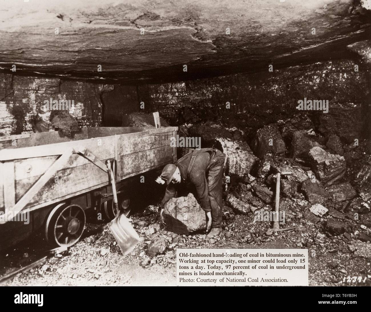 A miner, working at the end of a small, dark passage in a bituminous mine, bends down to lift a large piece of coal into an open rail car, 1920. Image courtesy National Coal Association/US Department of Energy. () Stock Photo