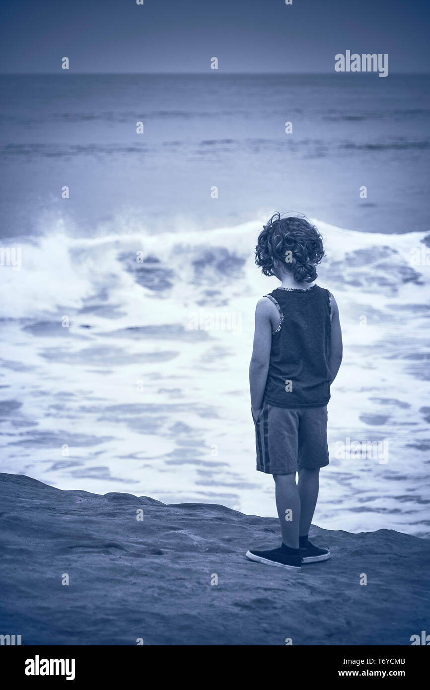 A young boy with curly hair wearing tanktop and shorts stands on the beach  and looks out to sea on overcast day in San Diego California Stock Photo -  Alamy