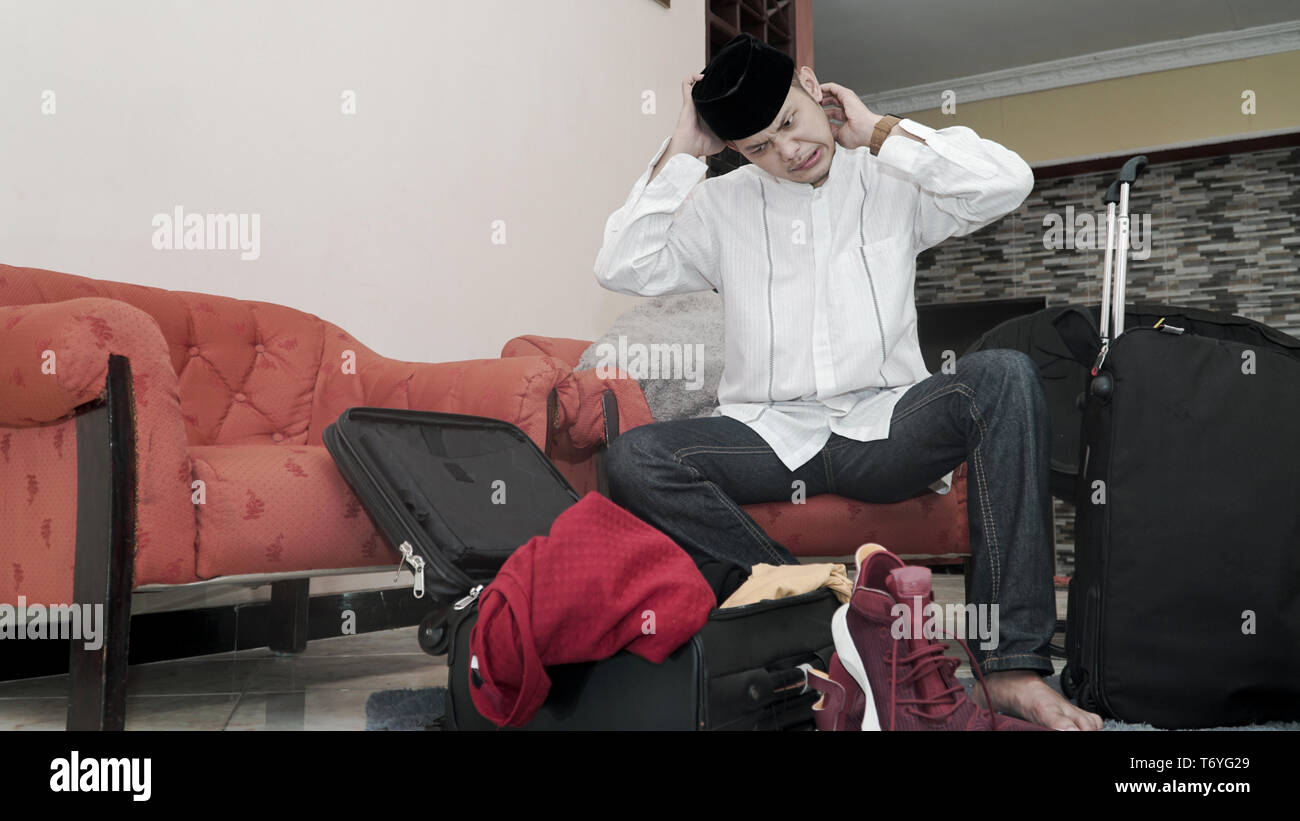 Muslim Asian man with songkok or head cap sitting on red couch stressed out prepare for traveling with stuff on black luggage suitcase -image Stock Photo