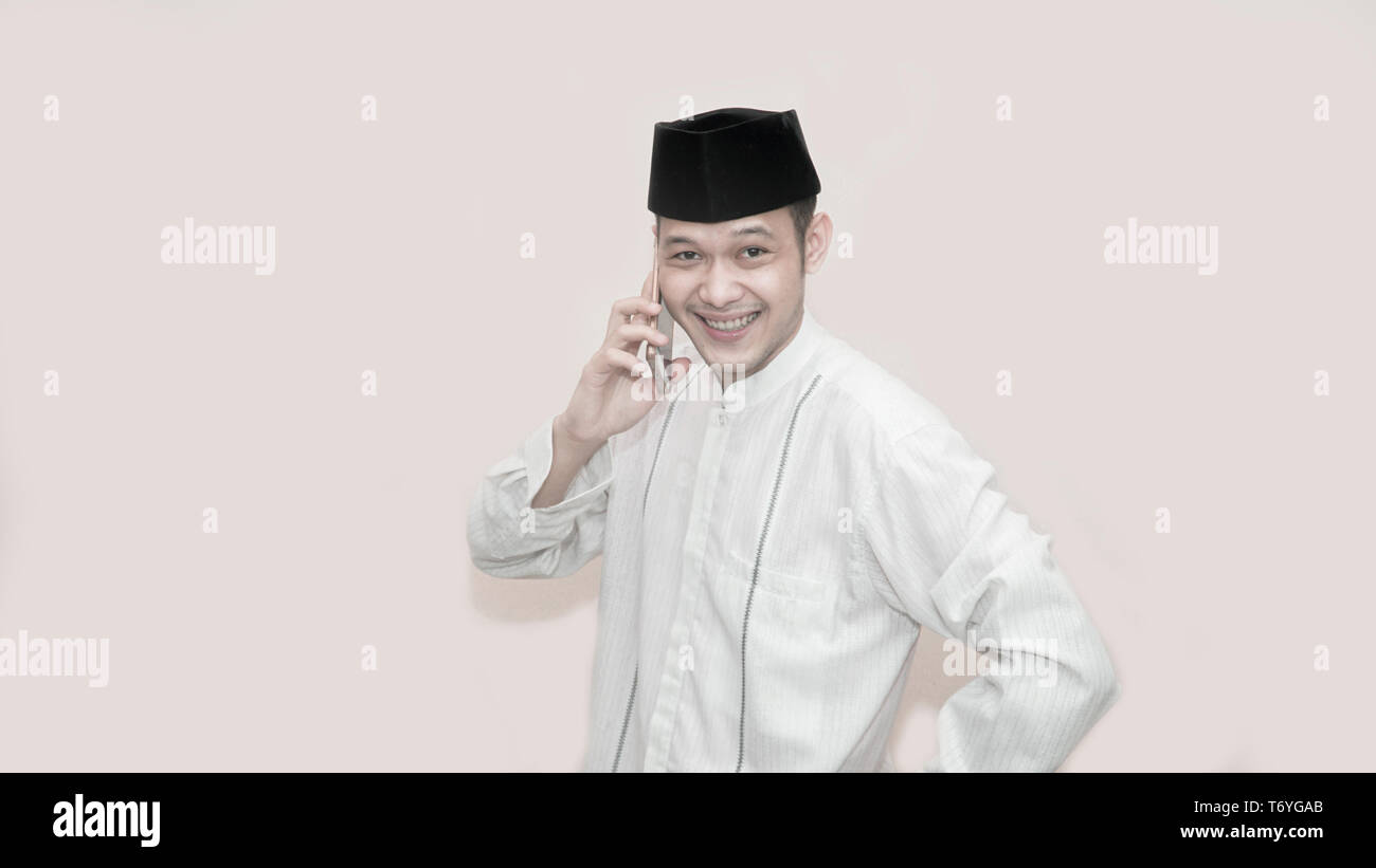 portrait of Funny an asian muslim man with head cap and baju koko taking a call for two way comunication -image Stock Photo