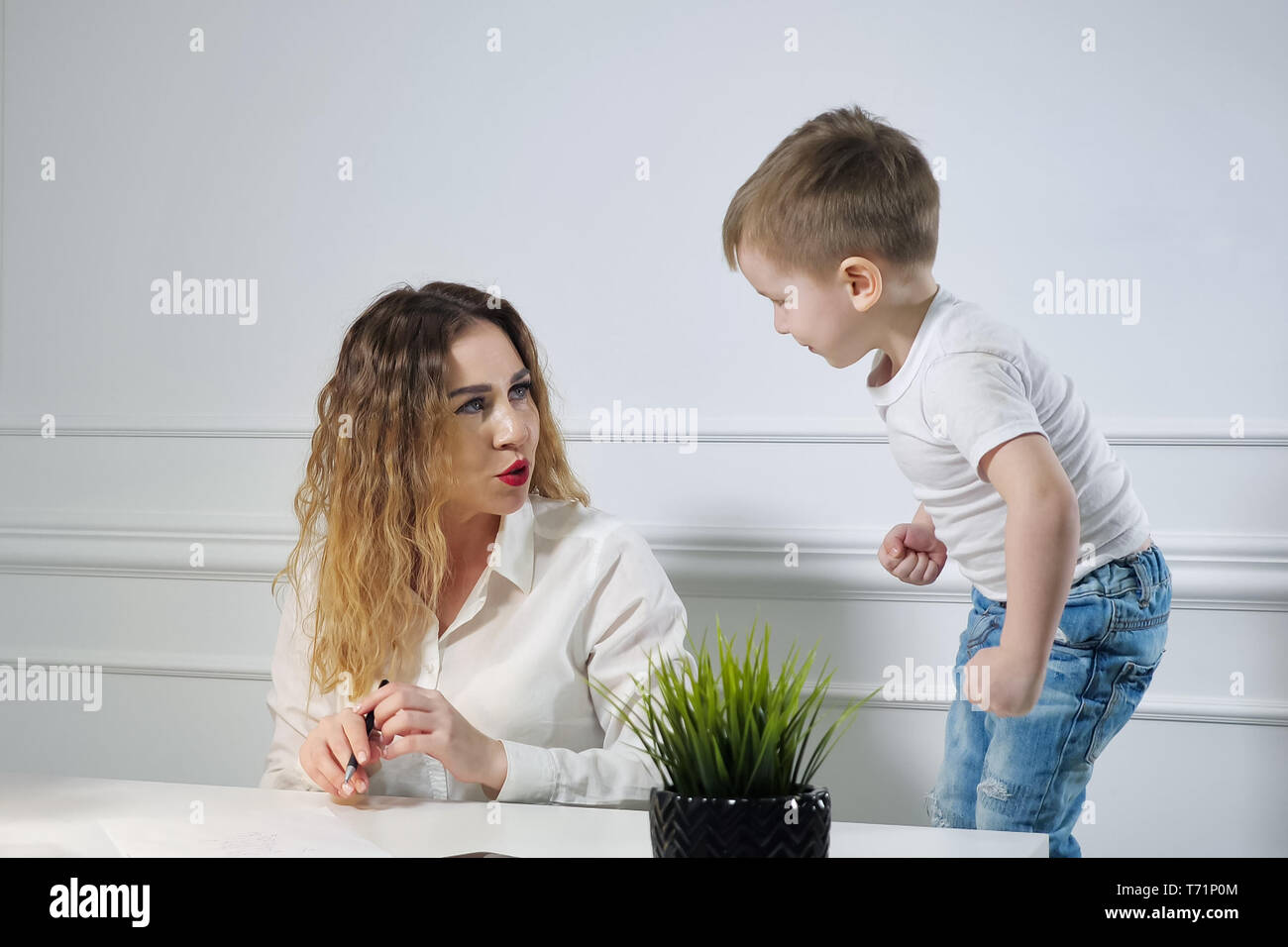 Young woman secretary with her little son in the workplace in office. Child requires his mother to play with him banging his fist on the table. Working mom. Stock Photo