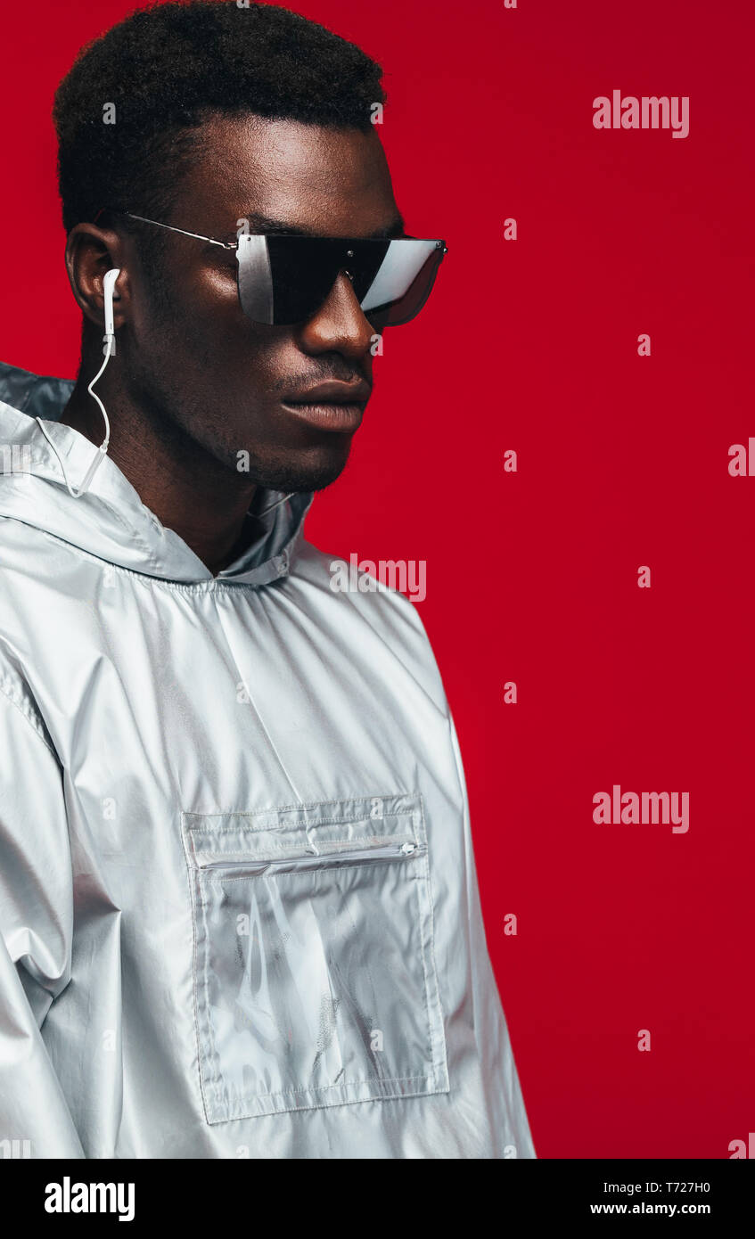 Trendy fashion portrait of man wearing silver outfit and stylish sunglasses.  Man in silver hooded shirt wearing earphones and sunglasses against red b  Stock Photo - Alamy