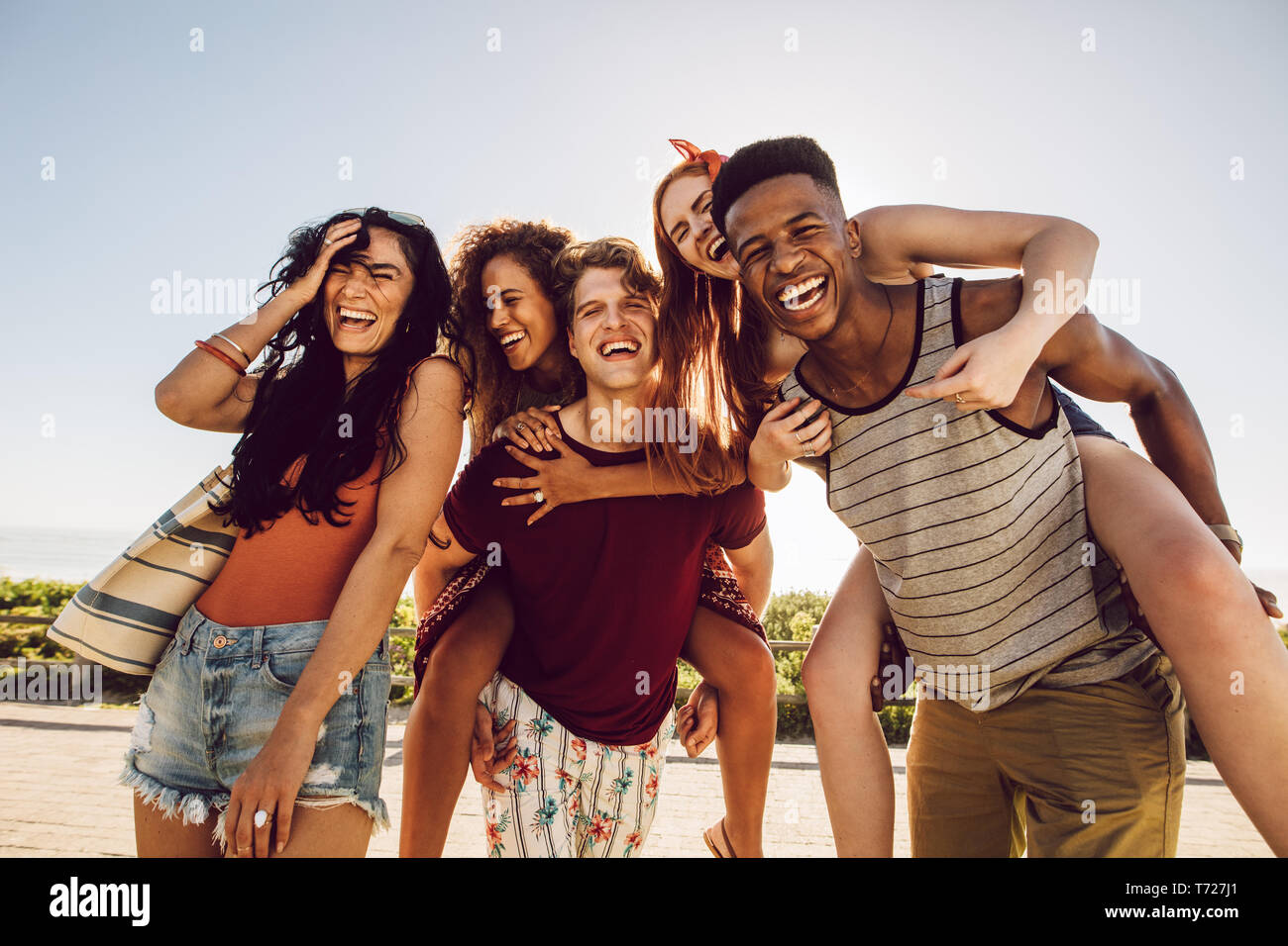 Group of happy friends having fun together outdoors. Men piggybacking female friends and laughing. Summer vacations. Stock Photo