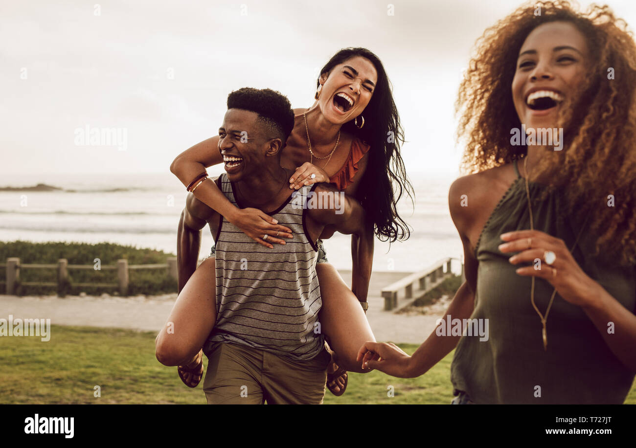 Excited young man carrying his girlfriend on his back with female friend walking in front. Diverse group of friends enjoying on a vacation. Stock Photo