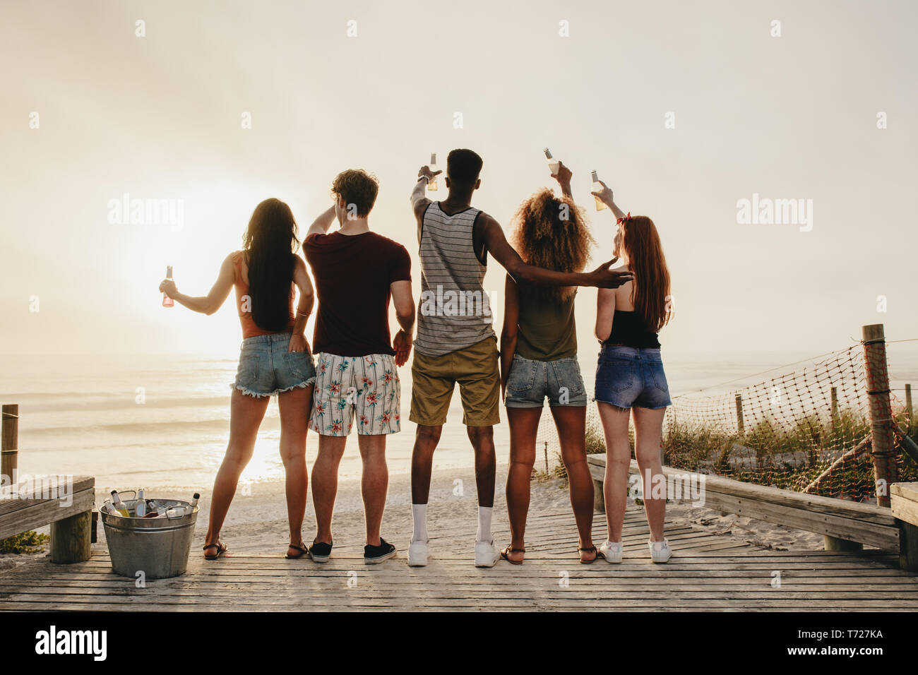 Rear view of young friends standing on boardwalk at the beach raising beer bottle at the sea. Group of friends having party at the beach. Stock Photo