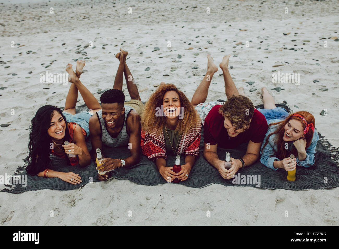 Cheerful young friends lying outdoors on a plaid with beer bottles. Young men and women laughing at the sea shore with beers. Stock Photo
