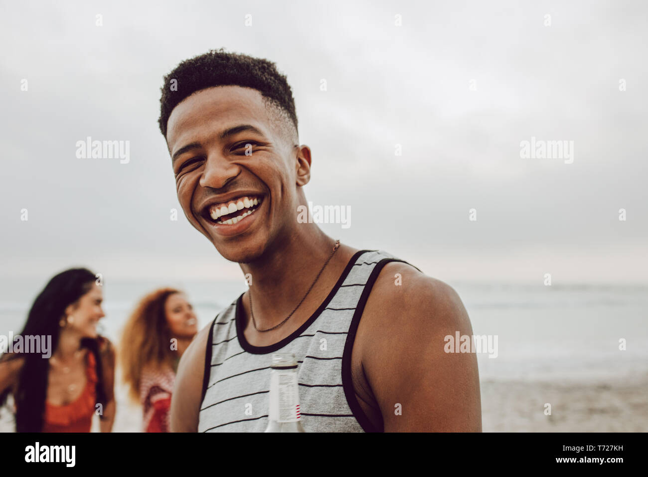 Handsome young african man on the beach with friends at the back. Smiling male standing at the beach with group of friends in background. Stock Photo
