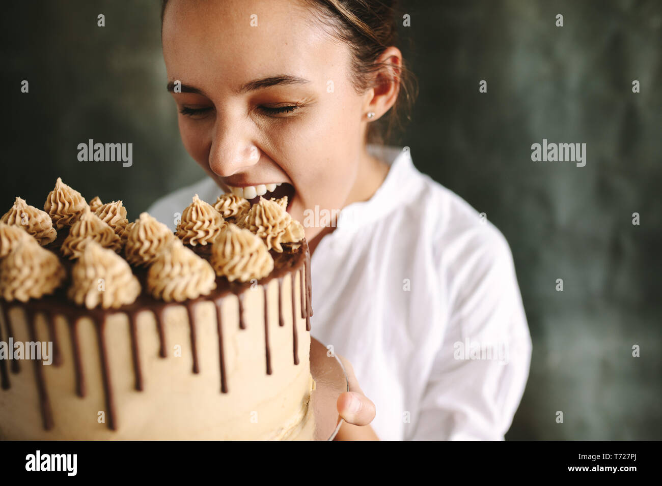 Female confectioner eating whole cake. Woman chef holding a big cake in hand and taking a bite. Stock Photo