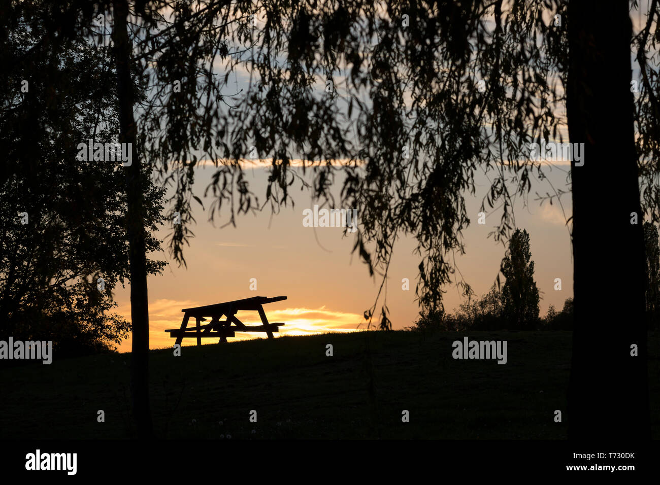 Beautiful UK sunset landscape photograph with view of isolated picnic table in silhouette on UK hillside as evening spring sun is setting in the sky. Stock Photo