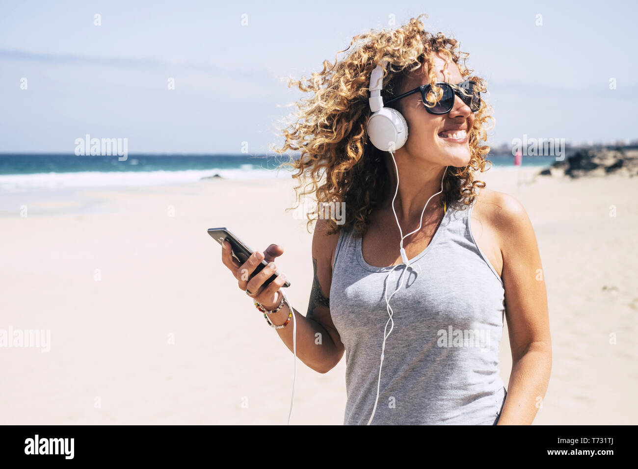 Cheerful beautiful young people blonde woman curly hair enjoying the summer holiday vacation at the beach listineg music from modern phone and headpho Stock Photo