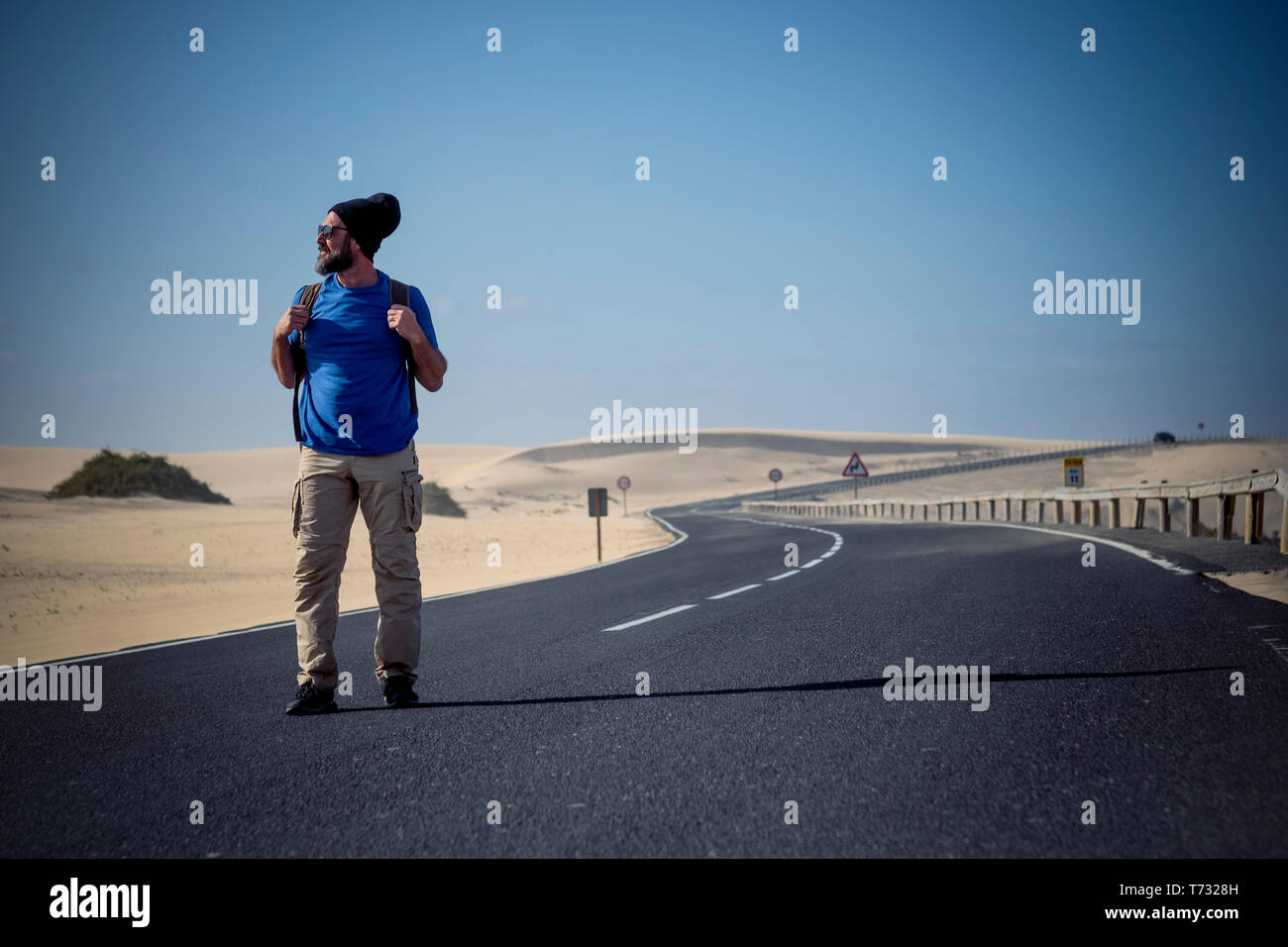 Coloured image of people man with travel backpack walking in the middle of the road with desert dunes around - alternative traveler concept for summer Stock Photo