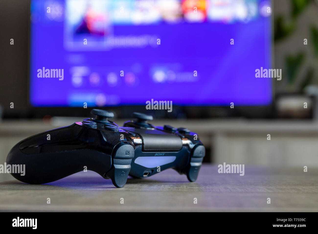 A portrait of a Playstation 4 controller, which is turned on, in front of a  blurred television showing the Playstation home screen Stock Photo - Alamy