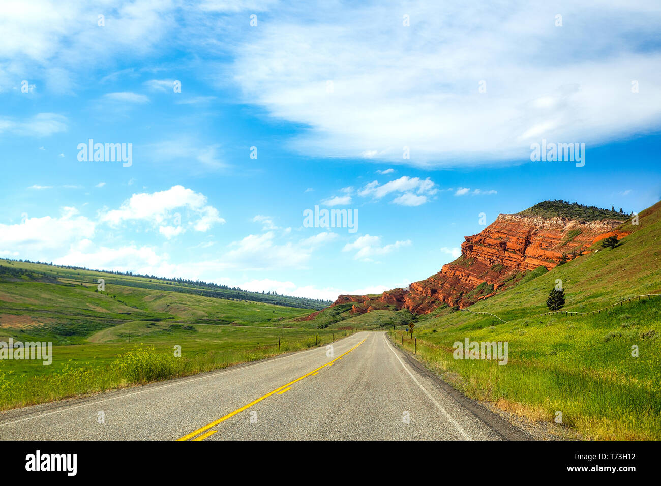 Tiered red rock formation jutting toward a winding highway between rolling hills along Chief Joseph scenic highway in a summer time Montana landscape Stock Photo